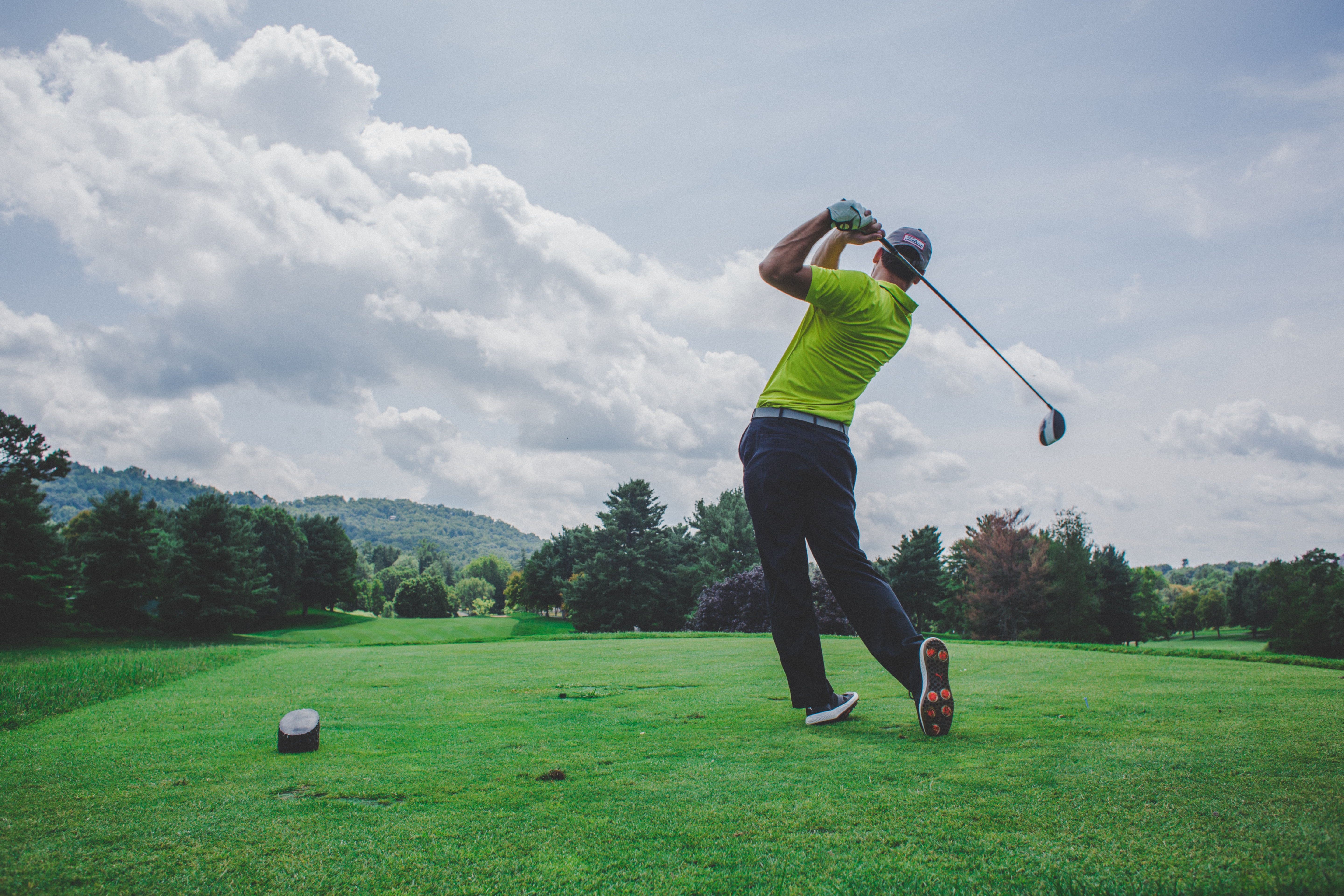 A man swings a golf club on a bright green, under a sky of scattered clouds.