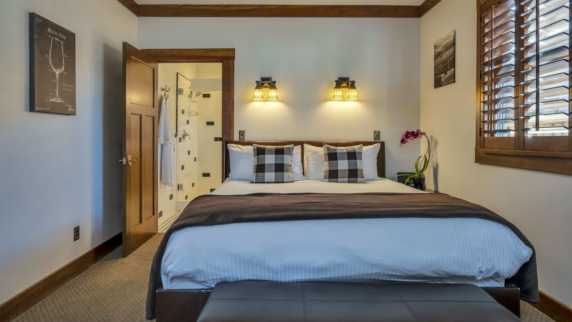 Bedroom with white walls, light colored carpeting, white bedding, leather bed bench, and view into bathroom