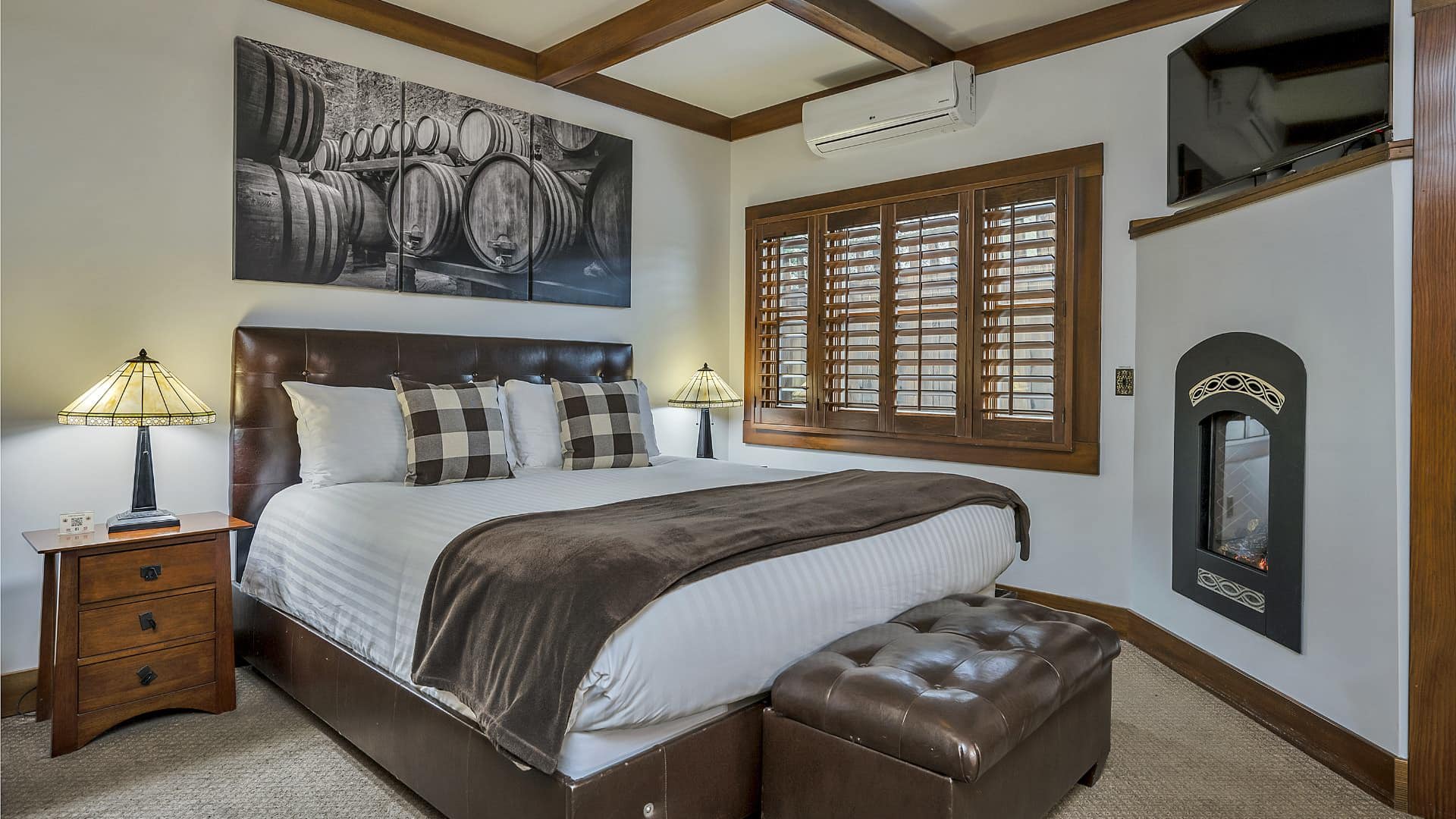 Bedroom with white walls, light colored carpeting, white bedding, leather bed bench, and electric fireplace