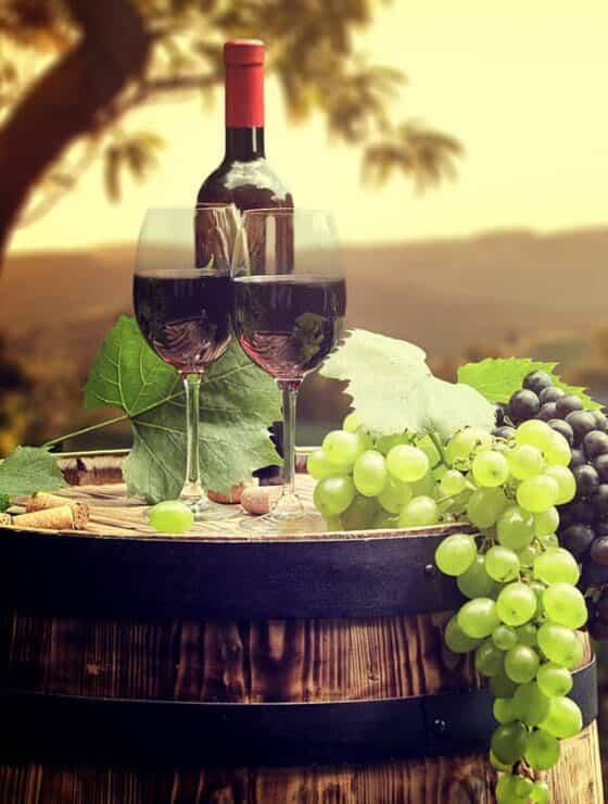 Two glasses full of red wine and green and red grapes sitting on top of a wooden barrel