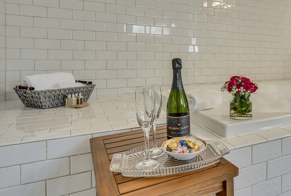 Champagne flutes, bottle of Champagne, and bowl of chocolates on glass tray sitting on wooden table next to a large soaker tub surrounded by white tile