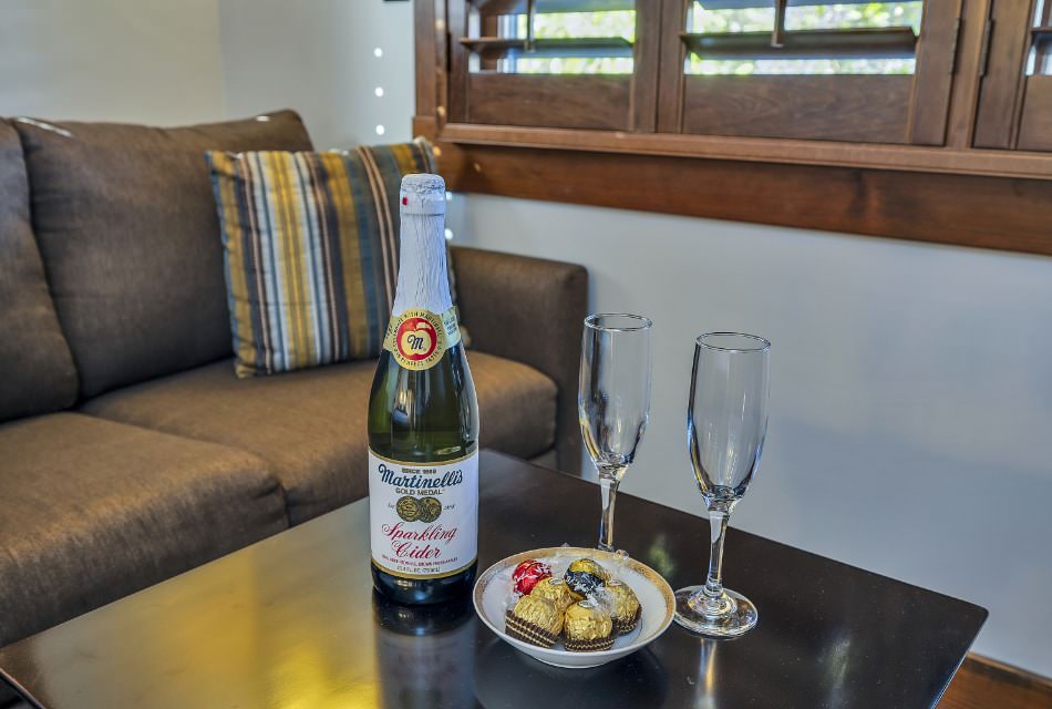 Close up view of a bottle of sparkling cider, two Champagne flutes, and bowl of chocolates on brown wooden end table
