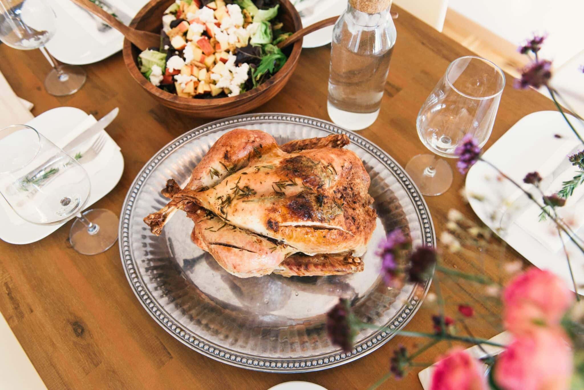 A table setting with a roasted chicken at the center and purple and pink flowers nearby.