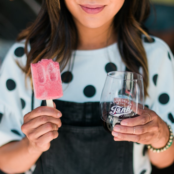 A woman in a black and white polka dot top holds a Tank Garage Winery wine glass in one hand and a pink popsicle in the other.