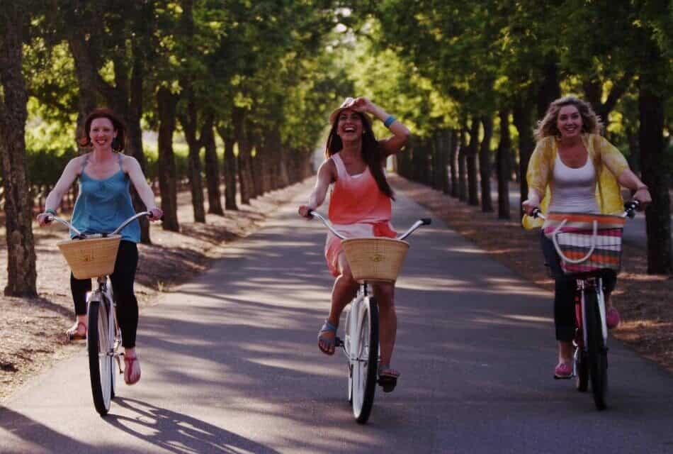 Three woman smiling and riding bicycles on paved road surrounded by trees