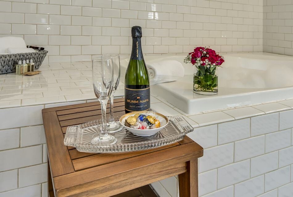 Champagne flutes, bottle of Champagne, and bowl of chocolates on glass tray sitting on wooden table next to a large soaker tub surrounded by white tile