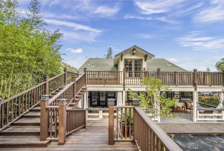 Exterior view of property painted white with gray trim, large two-story backyard patio with steps up to second story, and surrounded by green trees